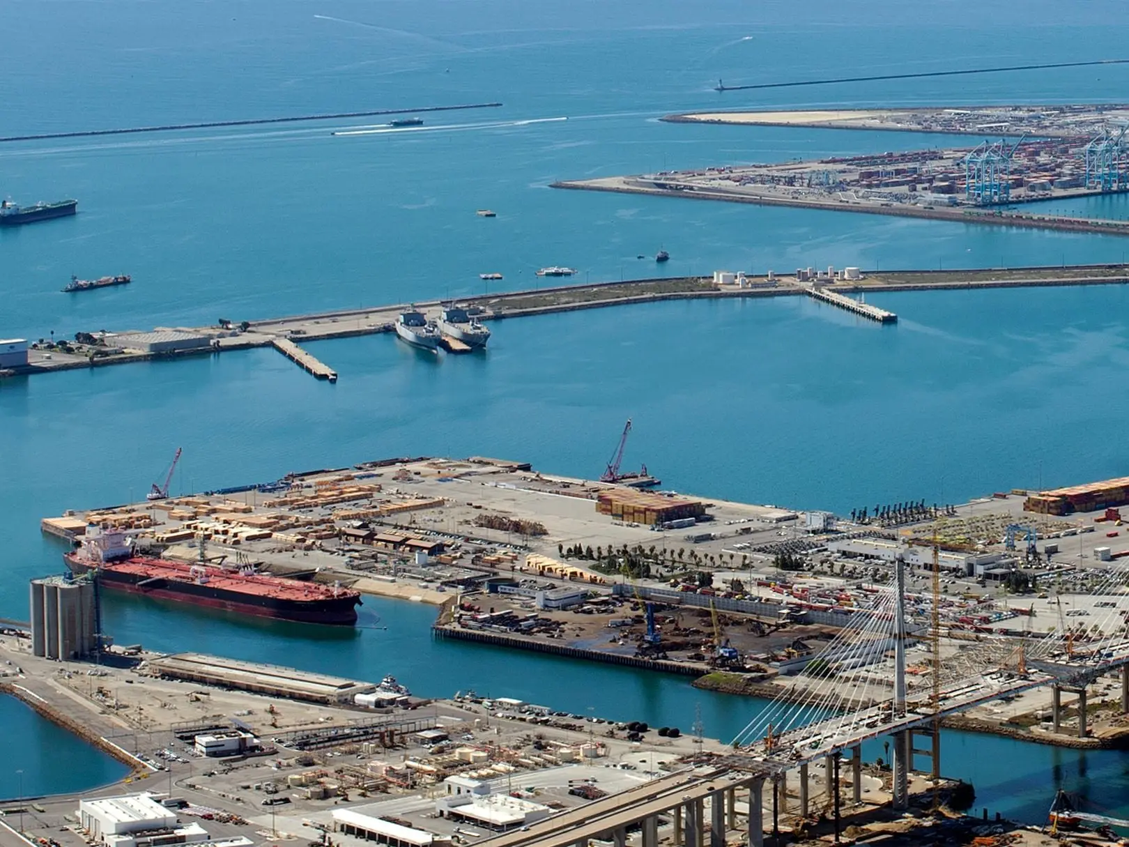 A sunlit harbour with a tanker at berth, island terminals and a cable-stayed road bridge, seen from the air.