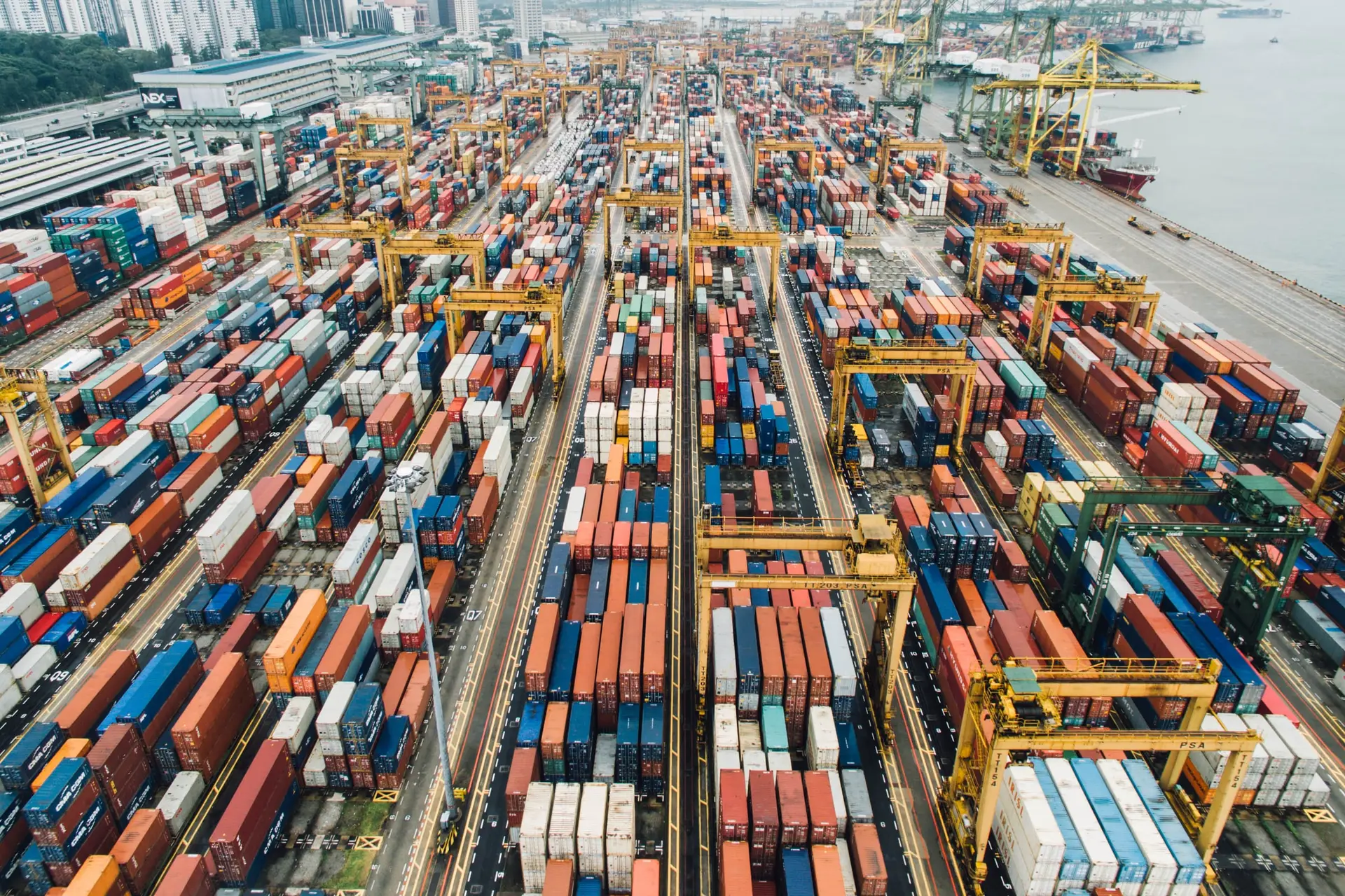 A container terminal from the air in bright daylight, with rows of colourful containers, yellow gantry cranes and a ship alongside the quay.