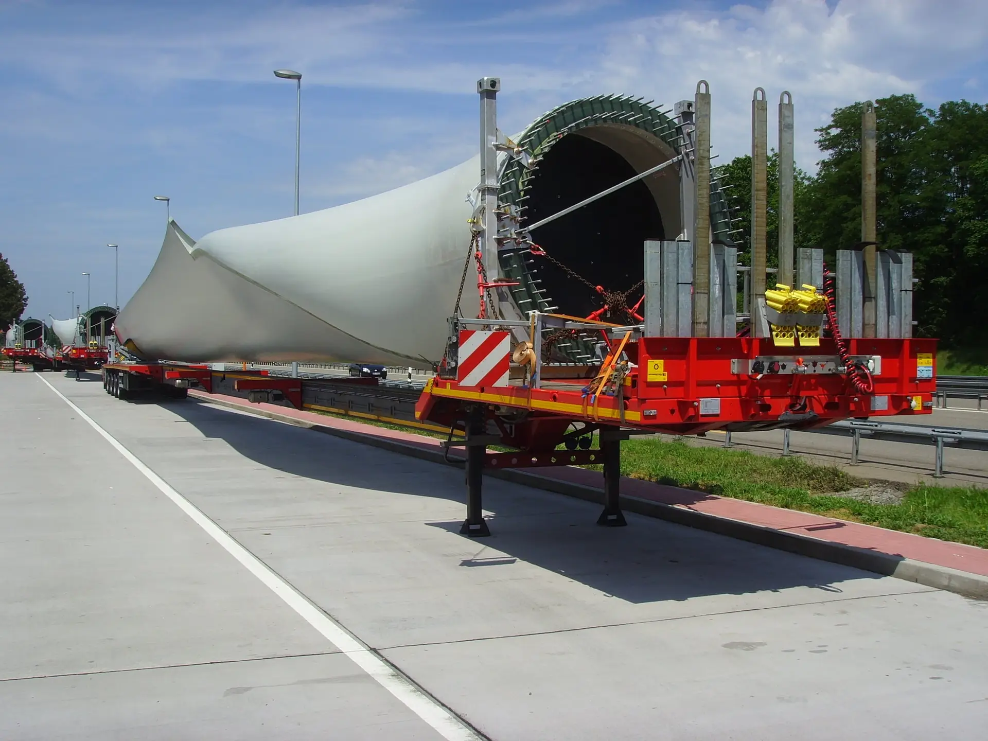 The root end of a wind turbine blade on a red extendable trailer at a motorway rest area, with more blade trailers lined up behind under a blue sky.