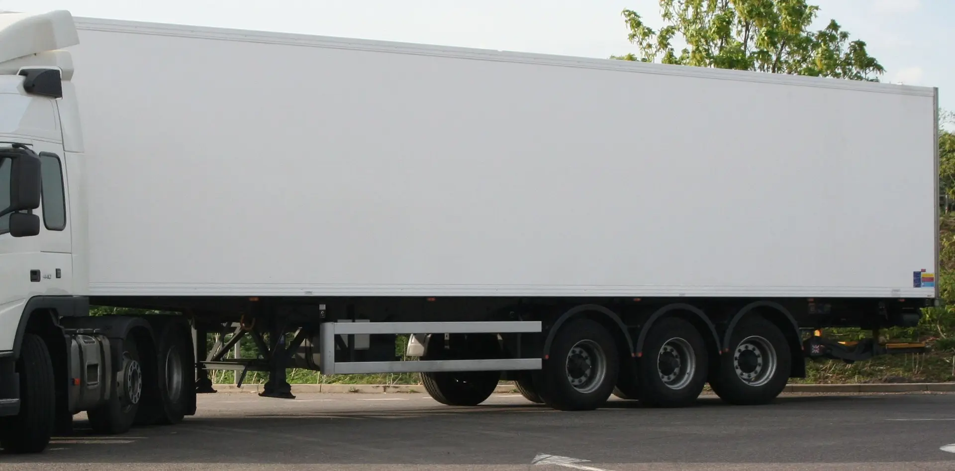 A plain white refrigerated semi-trailer coupled to a white tractor unit, parked in a yard with trees behind.