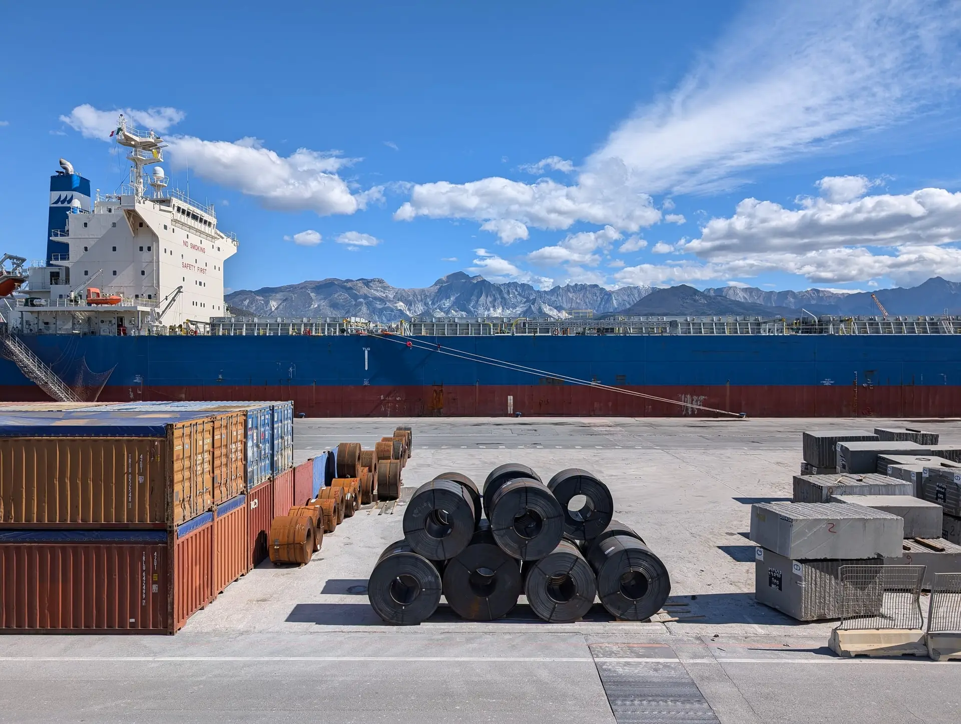 Steel coils and cargo staged on a quay beside a bulk carrier, mountains behind.