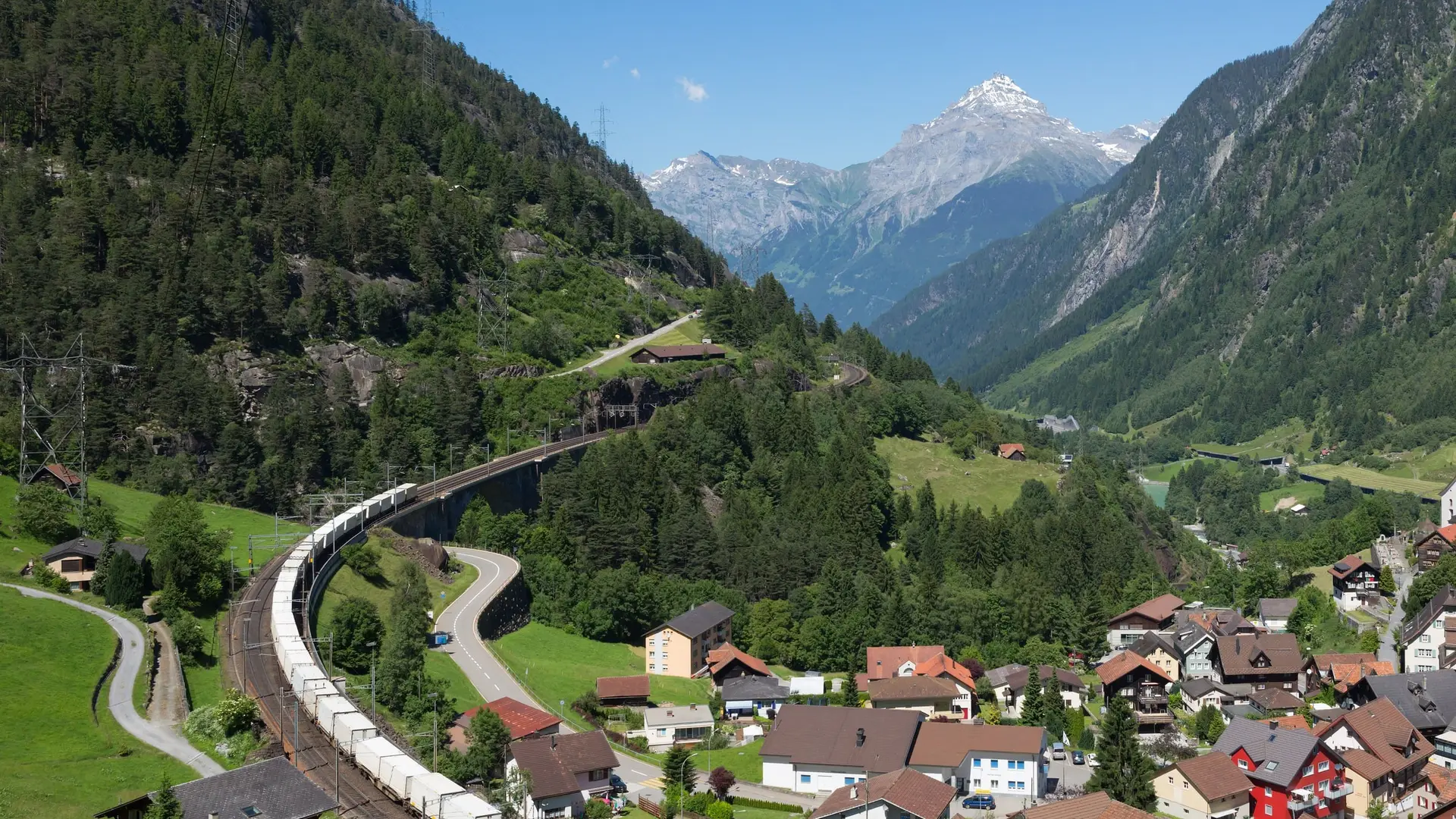 A container train of plain white boxes curving along a mountain railway above an Alpine village on a sunny day.