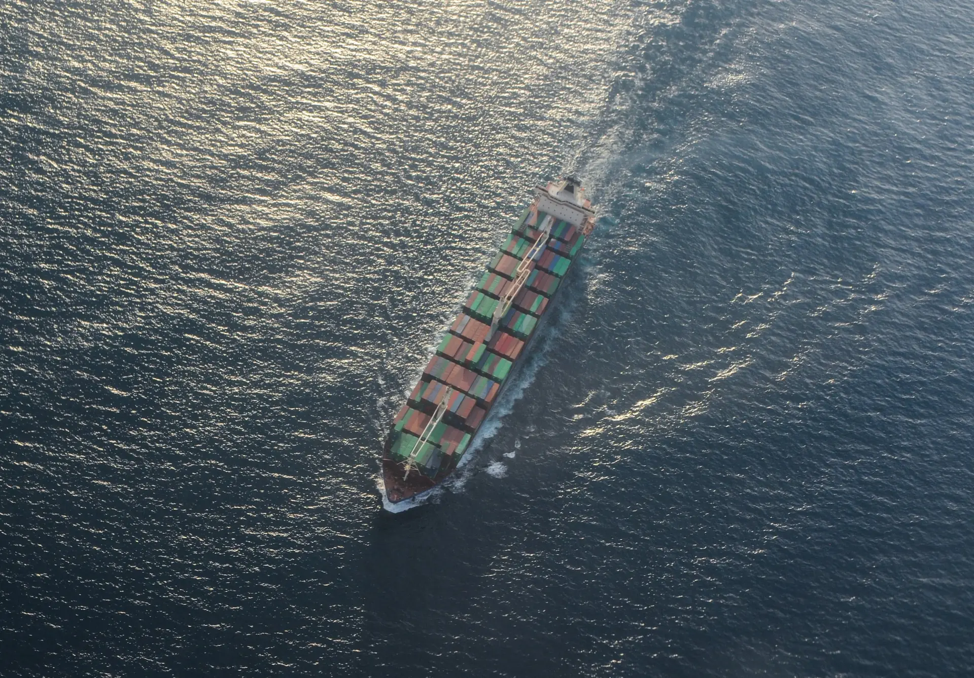 A loaded container ship under way on open sea, seen from directly above, with sunlight on the water.