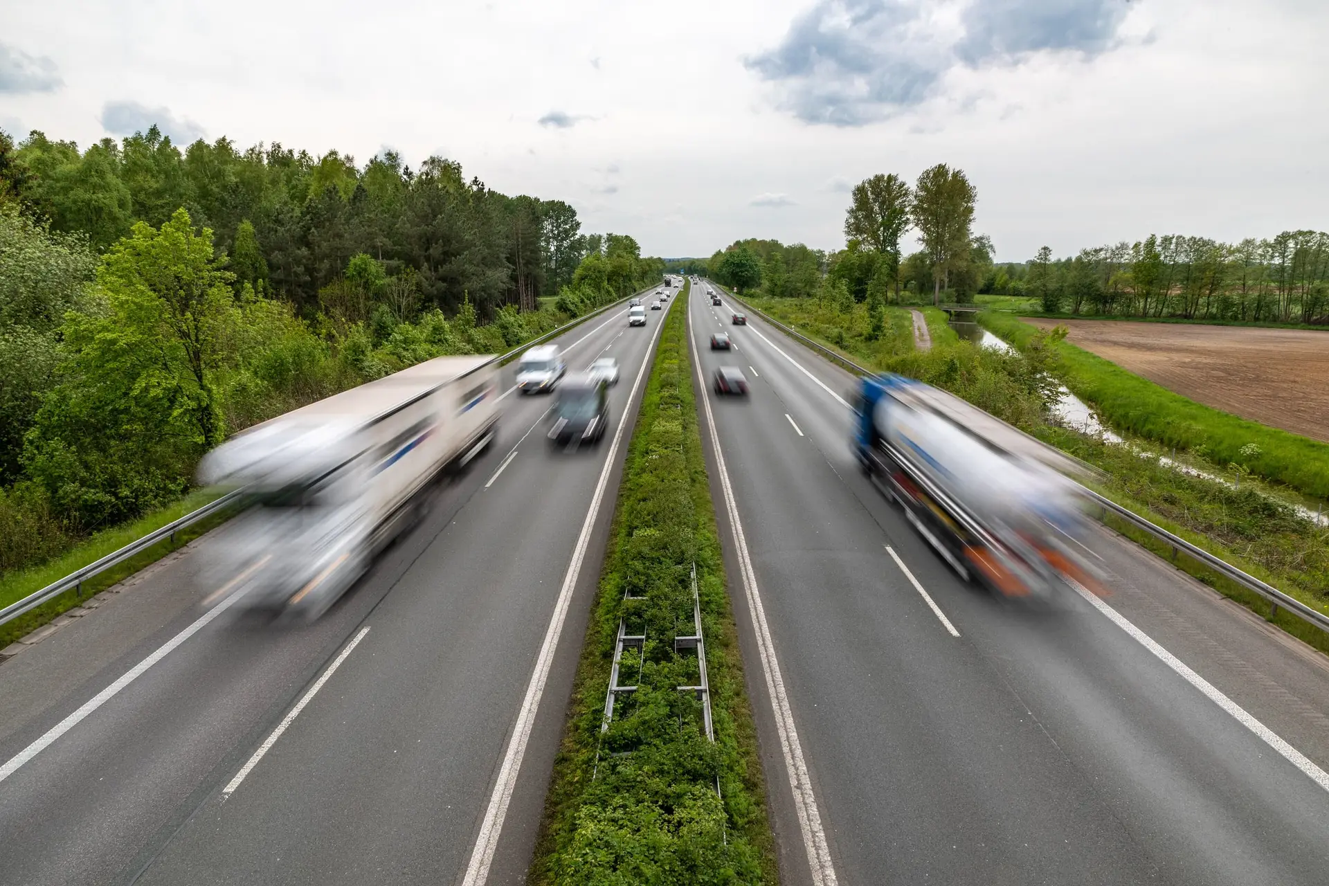 Lorries moving along a motorway between green trees and farmland, seen from a bridge on a bright day.