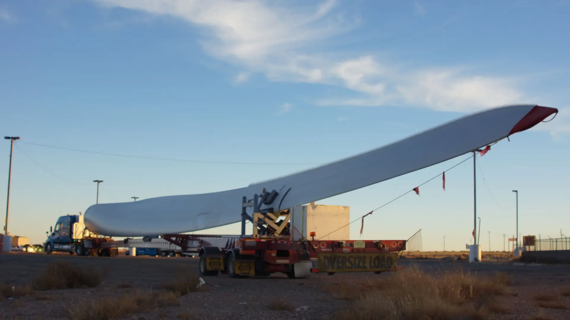 A wind turbine blade on a long multi-axle trailer with an oversize load banner, parked at a truck stop under a blue evening sky.