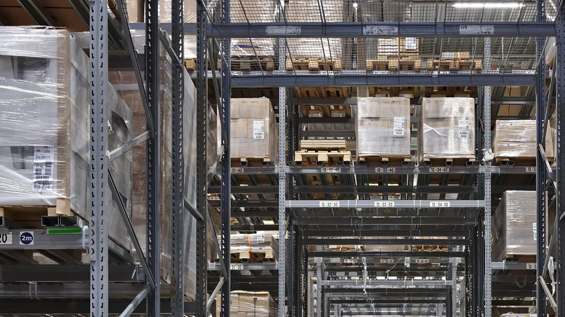Pallet racking stocked with shrink-wrapped goods along a long warehouse aisle, every bay labelled with its location.